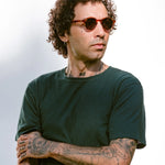 Man wearing sunglasses and a dark green t-shirt with tattoos on arms against a light background