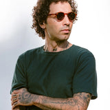Man wearing sunglasses and a dark green t-shirt with tattoos on arms against a light background